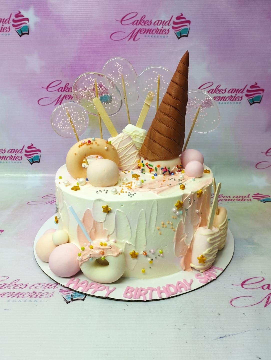 White 1-tier birthday cake with a large brown waffle cone topper, pastel donuts, popsicles, and clear isomalt lollipops with sprinkles.