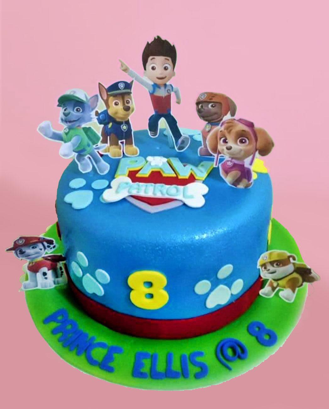 Blue Paw Patrol themed fondant cake with character printouts, paw prints, and a green fondant base board with a birthday message.