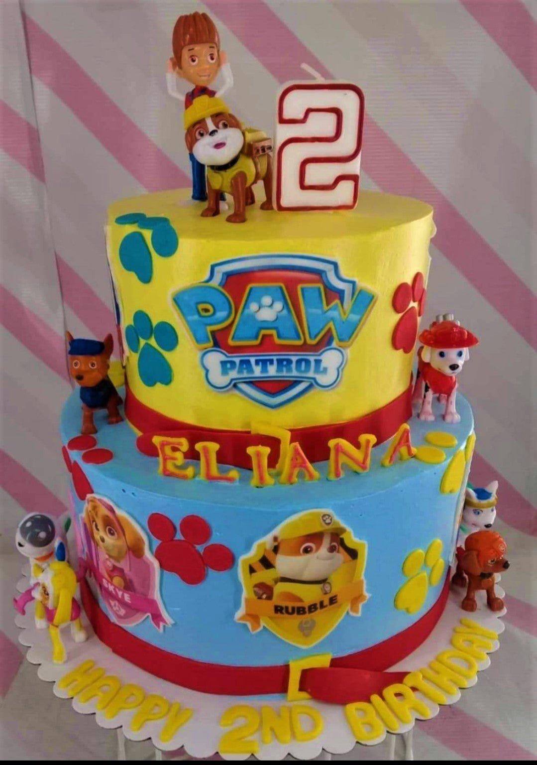 Two-tier Paw Patrol birthday cake with yellow and blue icing, character figurines, and a large number 2 candle.