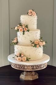 Elegant three-tier white wedding cake decorated with clusters of peach roses and delicate white icing floral textures on a gold stand.