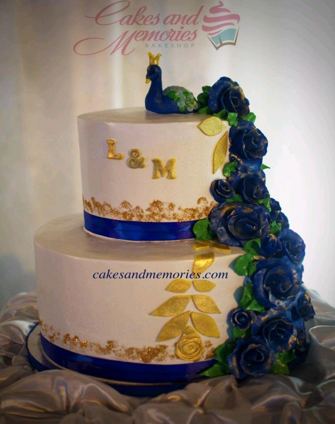 Elegant 2-tier white cake with royal blue roses, gold leaf accents, and a 3D peacock topper with a crown.