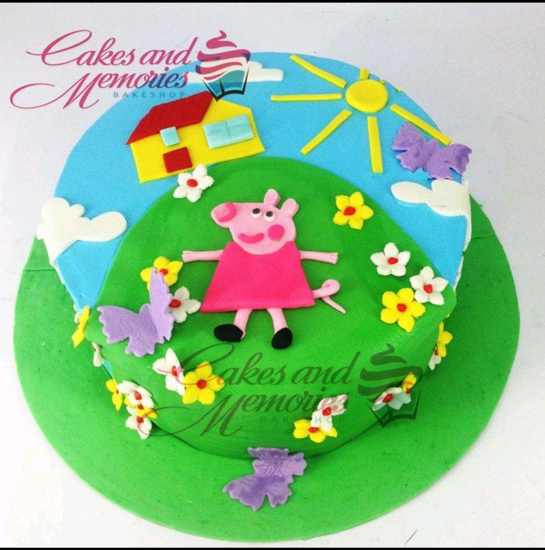 Peppa Pig themed fondant cake featuring a green hill, blue sky, sun, house, and Peppa Pig character with flowers and butterflies.