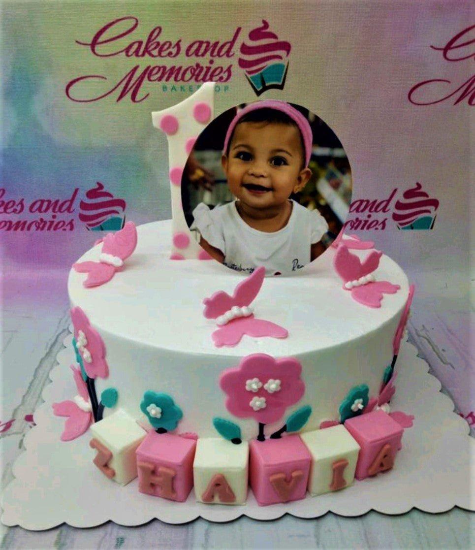White 1-tier birthday cake with a photo printout of a baby girl, pink fondant butterflies, flowers, and name blocks for Zhavia.