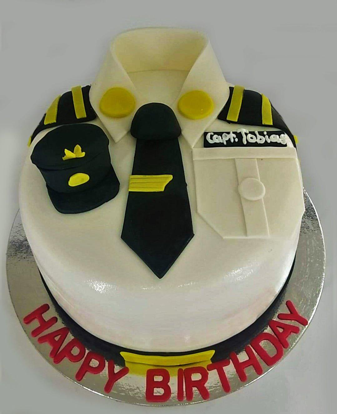 White pilot uniform fondant cake with black tie, pilot hat, and captain name tag for a birthday celebration.
