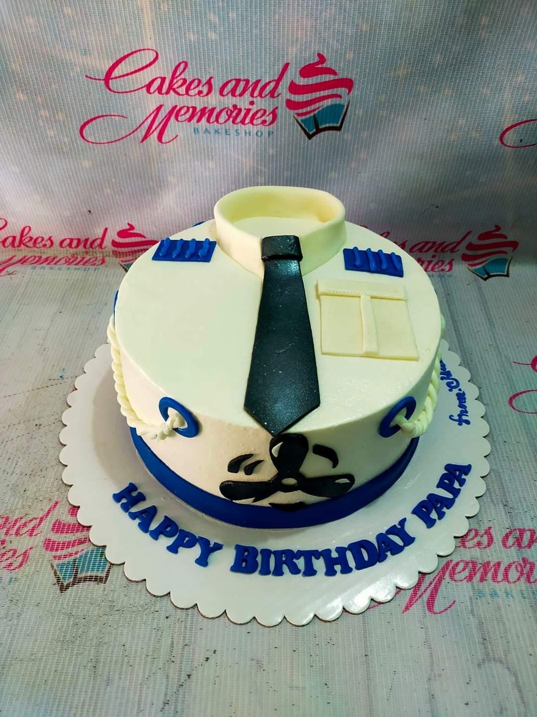 White 1-tier pilot uniform cake with a black tie, blue epaulettes, and a propeller design on the side.