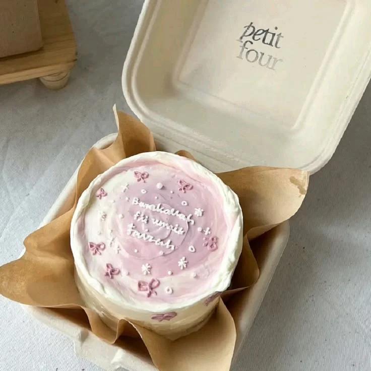 Pink and white bento cake with a textured palette knife finish, pink icing bows, and white icing stars in a takeout box.