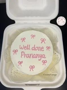 White bento cake with pink piped icing bows and a 'Well done Prananya' message in pink icing.