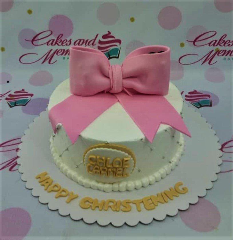 White 1-tier christening cake with a large pink fondant bow, silver dragees, and gold lettering on a scalloped base board.