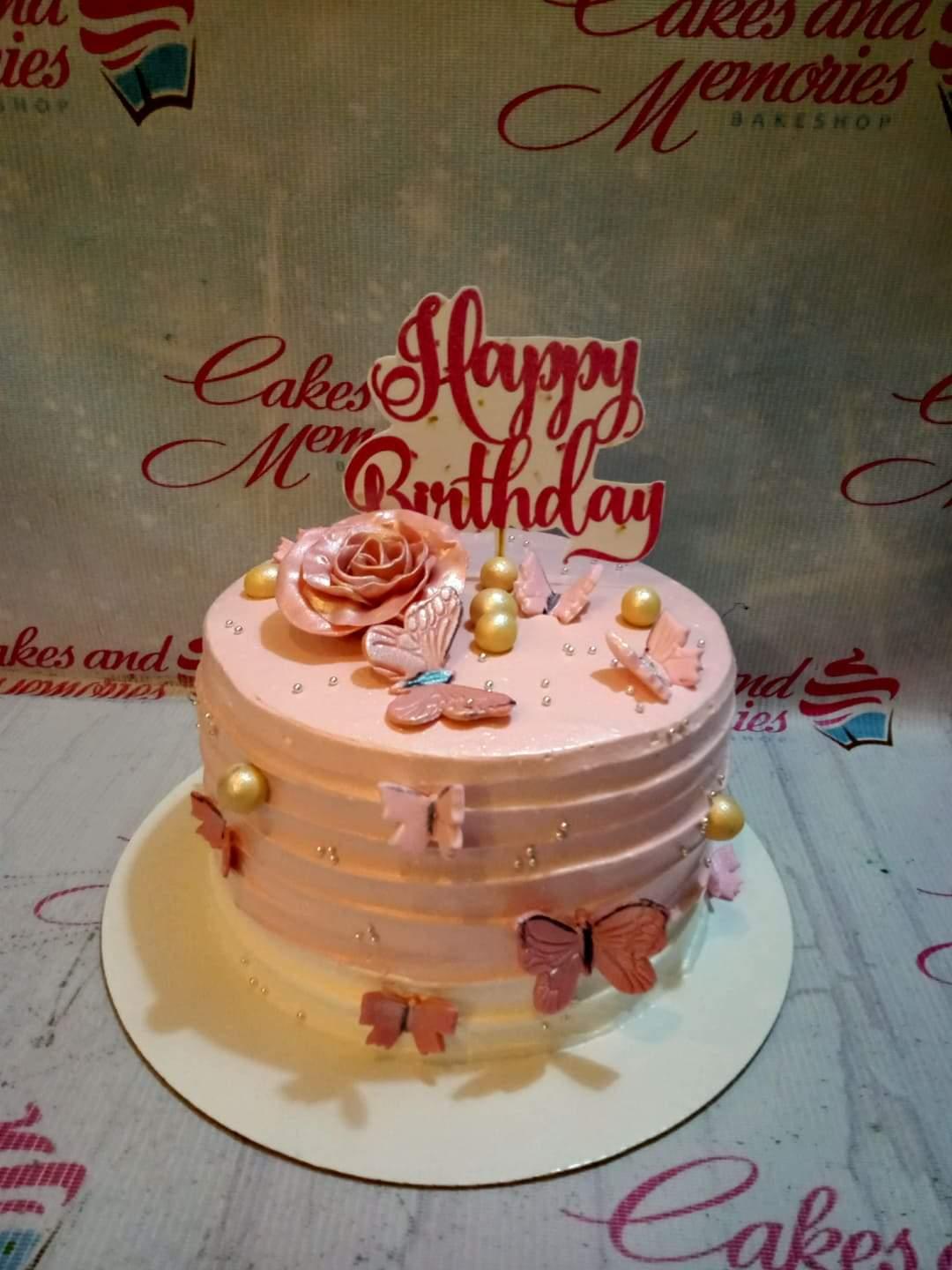 Pink soft-icing cake with a large rose gold rose, pink butterflies, gold spheres, and a Happy Birthday topper.