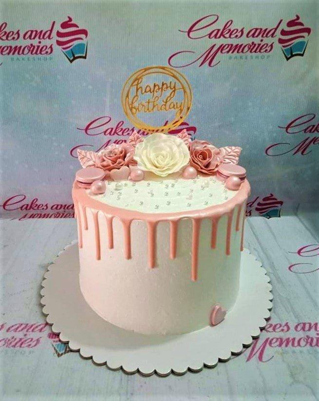 Elegant white cake with pink drip, pink and white edible roses, pink macarons, and a gold Happy Birthday topper.