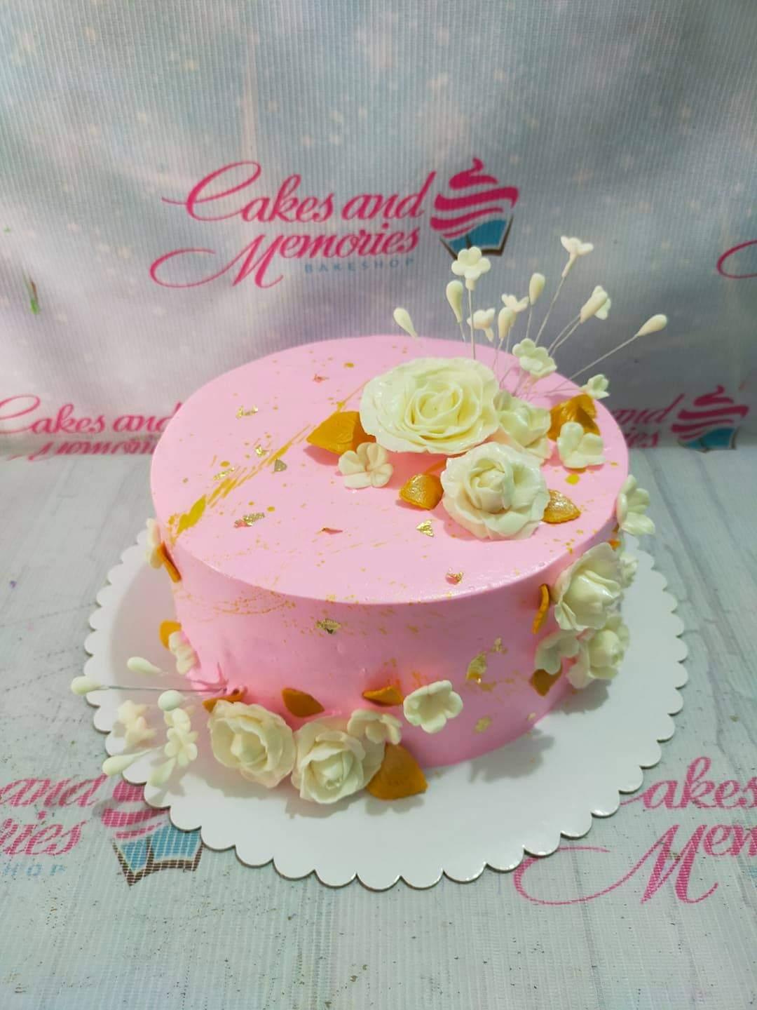Pink round cake decorated with white gumpaste roses, gold leaf accents, and small white blossoms on the top and sides.