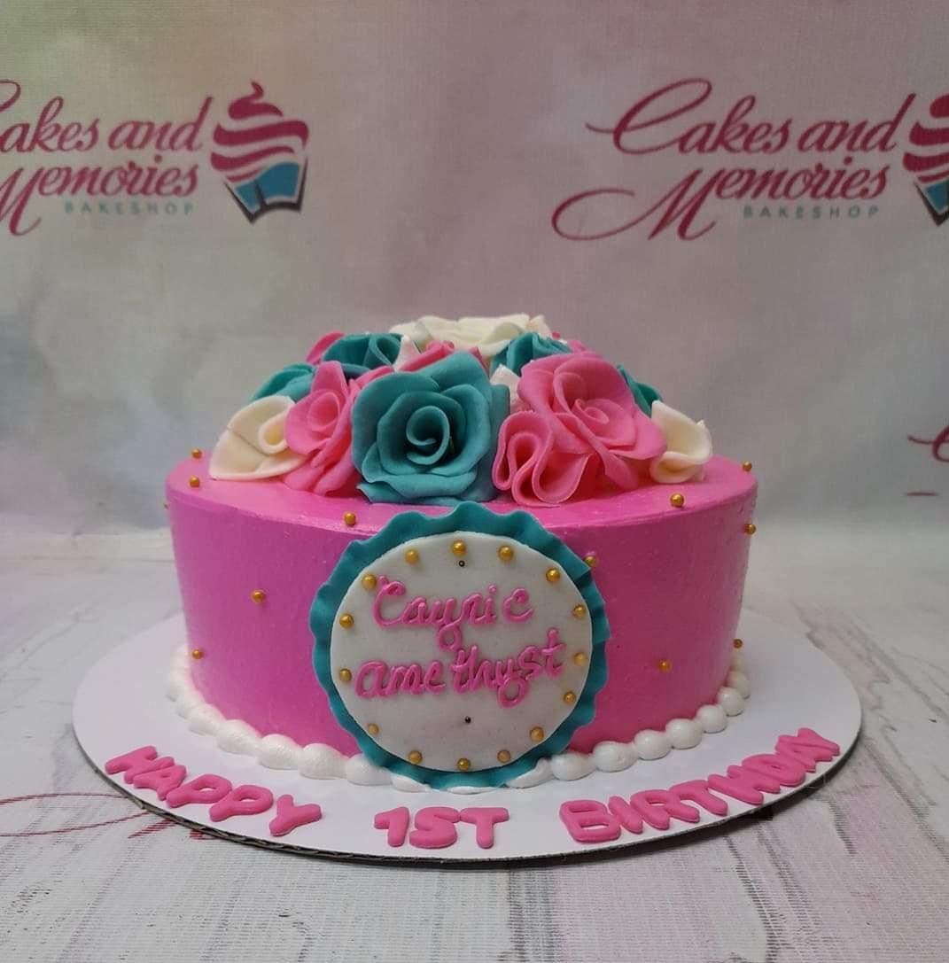 Pink round cake with teal, pink, and white fondant roses on top and gold dragees on the sides.
