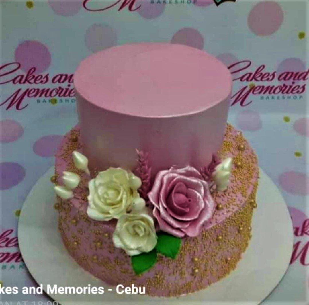 Two-tier pink cake decorated with large pink and white gumpaste roses and gold dragees on a pink textured base.