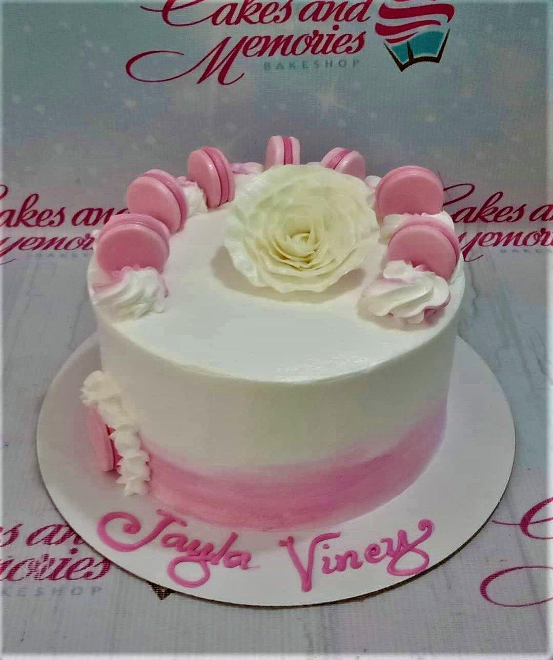 White and pink soft icing cake with pink macarons and a large white edible rose topper.