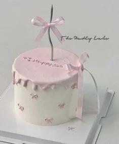 Minimalist white bento cake with pink icing drip, piped pink bows, and a tall silver candle with a pink ribbon.