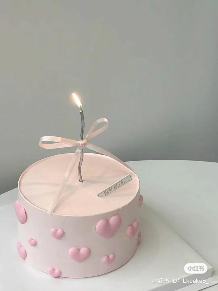 Minimalist pink cake with a single tall silver candle, a pink ribbon bow, and raised pink fondant hearts on the sides.