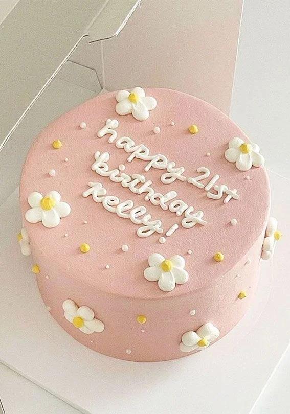 Minimalist pink birthday cake with white icing daisies and yellow centers, featuring white piped icing text for Kelly's 21st birthday.