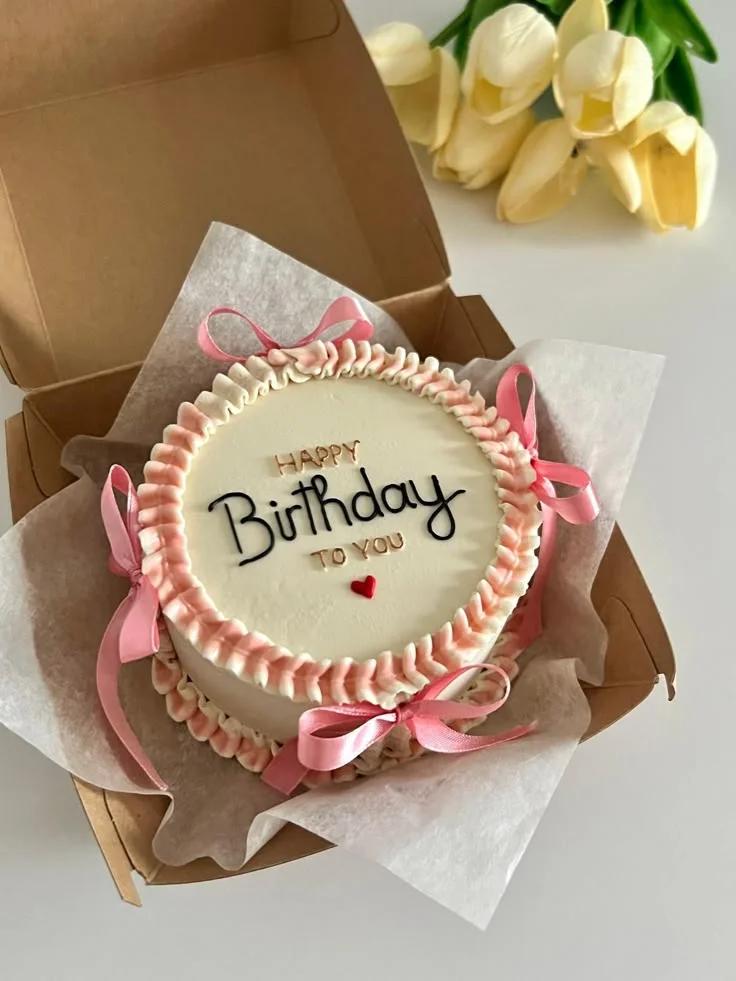 Minimalist white bento cake with pink ruffled icing borders, pink ribbon bows, and a Happy Birthday message in black and gold.