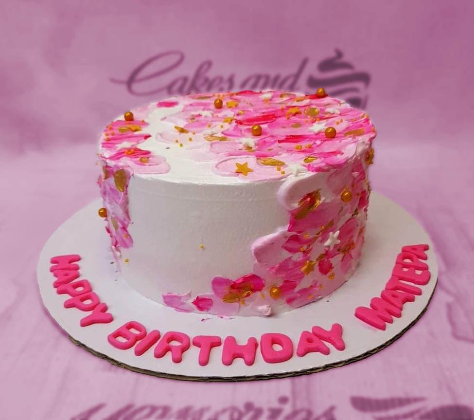 White round cake with pink palette knife icing strokes, gold dragees, and pink gumpaste birthday message on the board.
