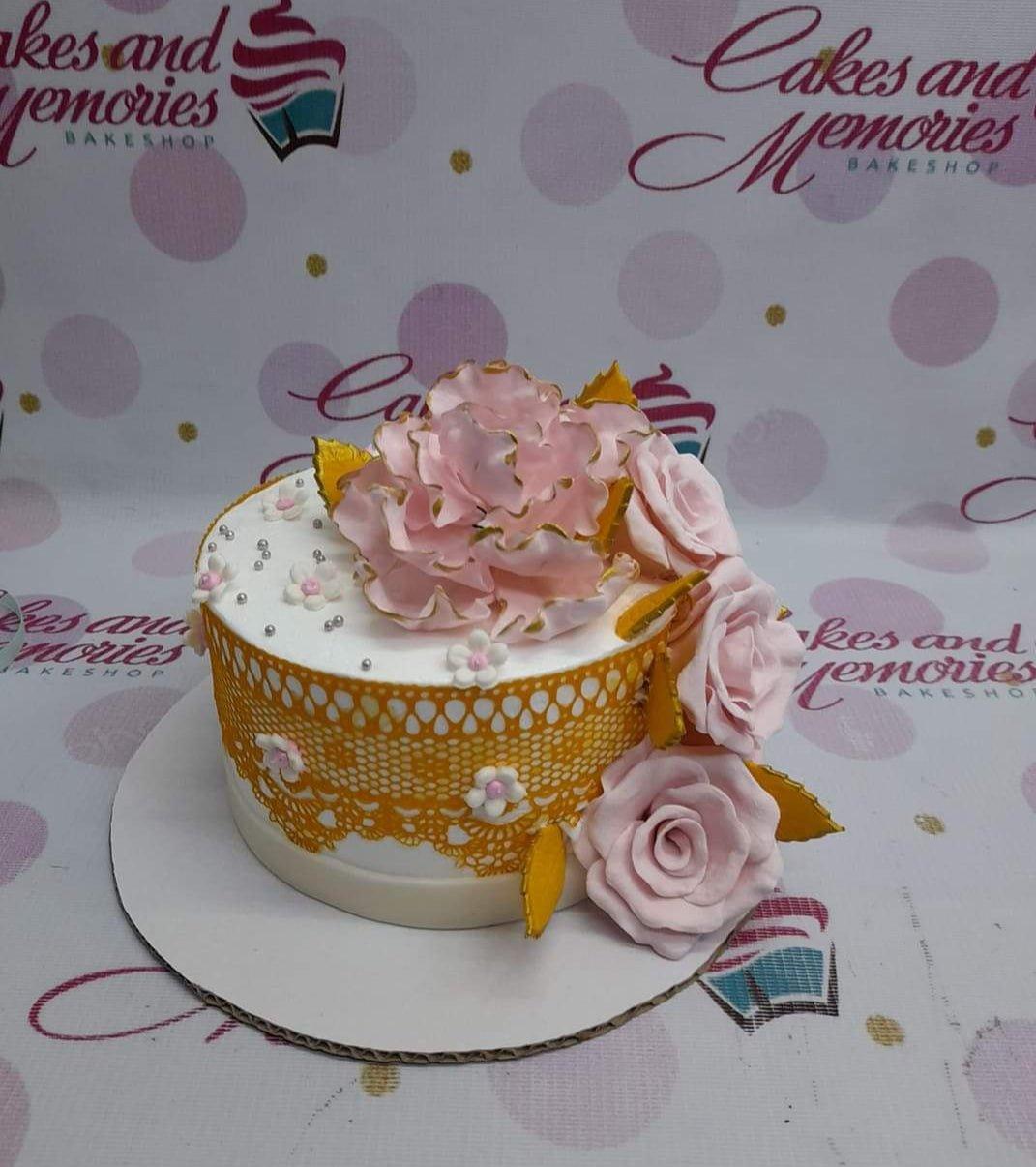 Elegant white fondant cake with a large pink gumpaste peony, gold lace wrap, and silver dragees.