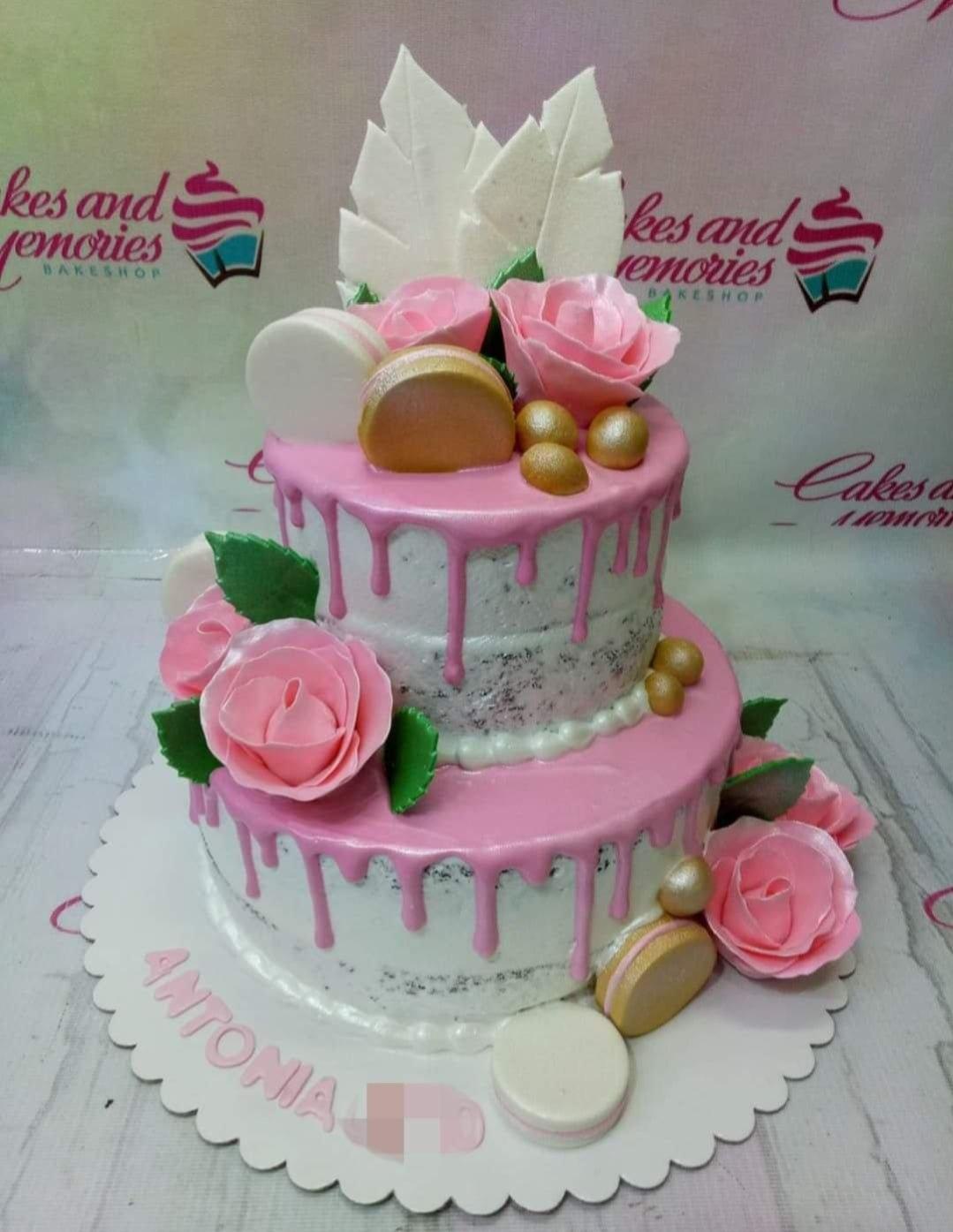 Two-tier semi-naked cake with pink icing drips, pink fondant roses, gold macarons, and gold spheres.