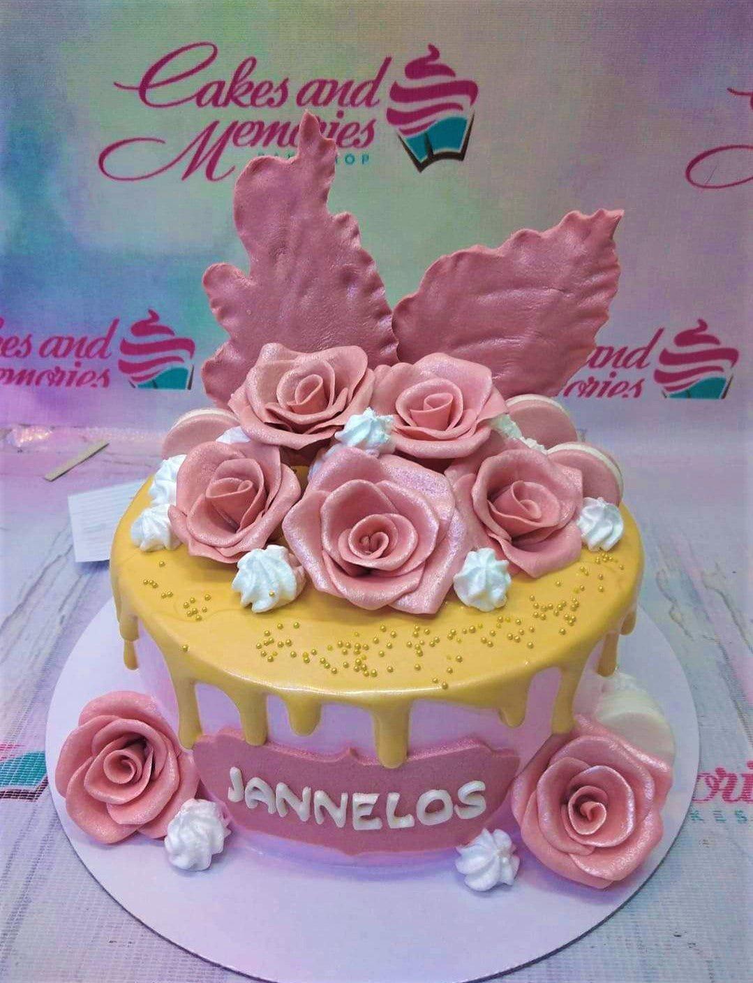 Pink and yellow drip cake with large pink gumpaste roses, white icing rosettes, and gold dragees.