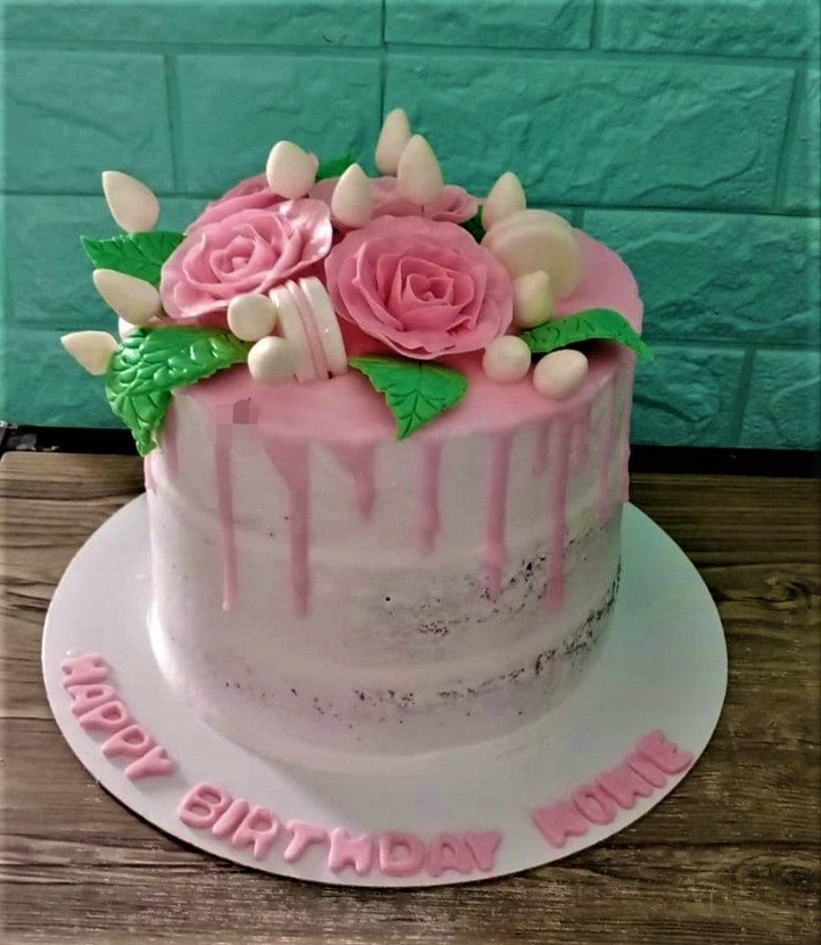 Pink semi-naked cake with pink fondant roses, green leaves, white macarons, and pink icing drips on a white board with a birthday message.