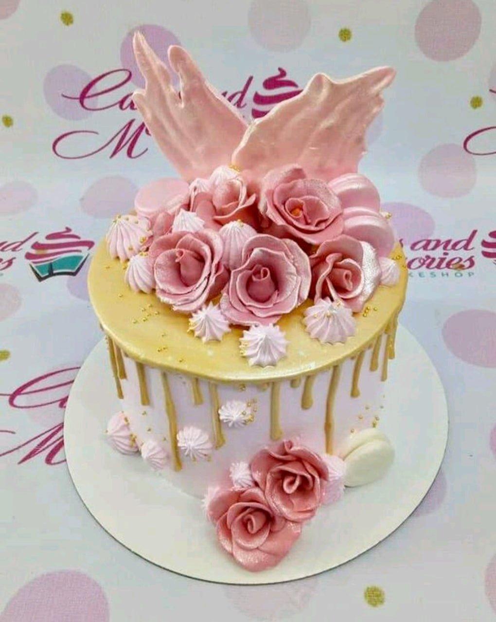 Pink and gold drip cake with pink fondant roses, meringue kisses, and large pink gumpaste wings on top.
