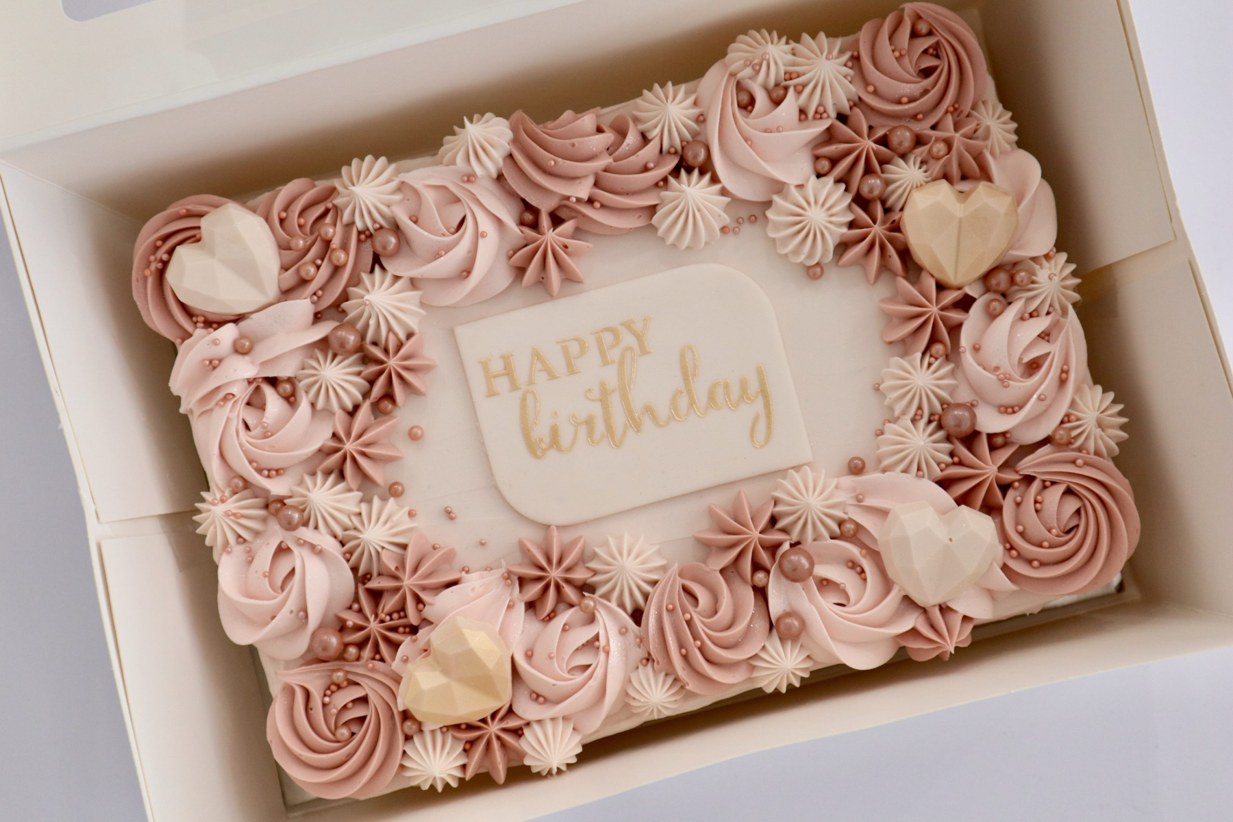 Rectangular soft icing cake with intricate pink and tan piped rosettes, geometric chocolate hearts, and a Happy Birthday message plaque.