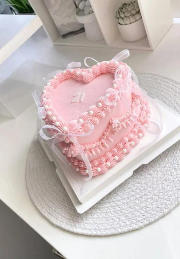 Heart-shaped pink vintage cake with intricate icing ruffles, white pearls, and delicate white ribbons.