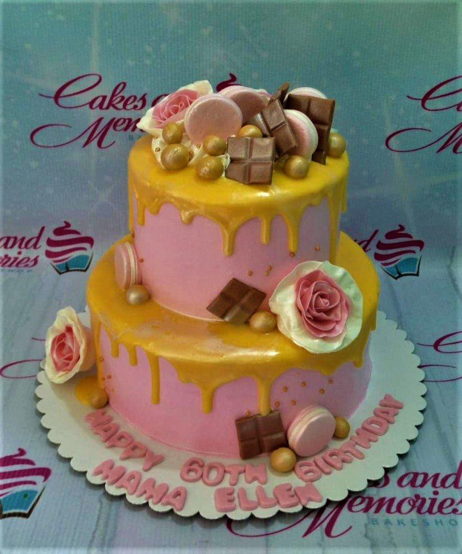 Two-tier pink birthday cake with yellow drip, pink macarons, chocolate bars, gold spheres, and pink icing roses.
