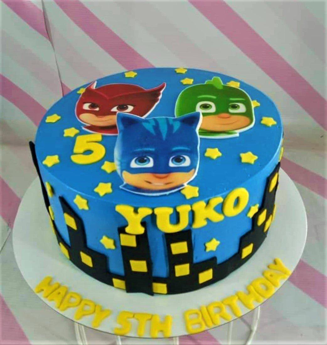 Blue PJ Masks themed birthday cake with Catboy, Owlette, and Gekko printouts, yellow stars, and a black city skyline silhouette.