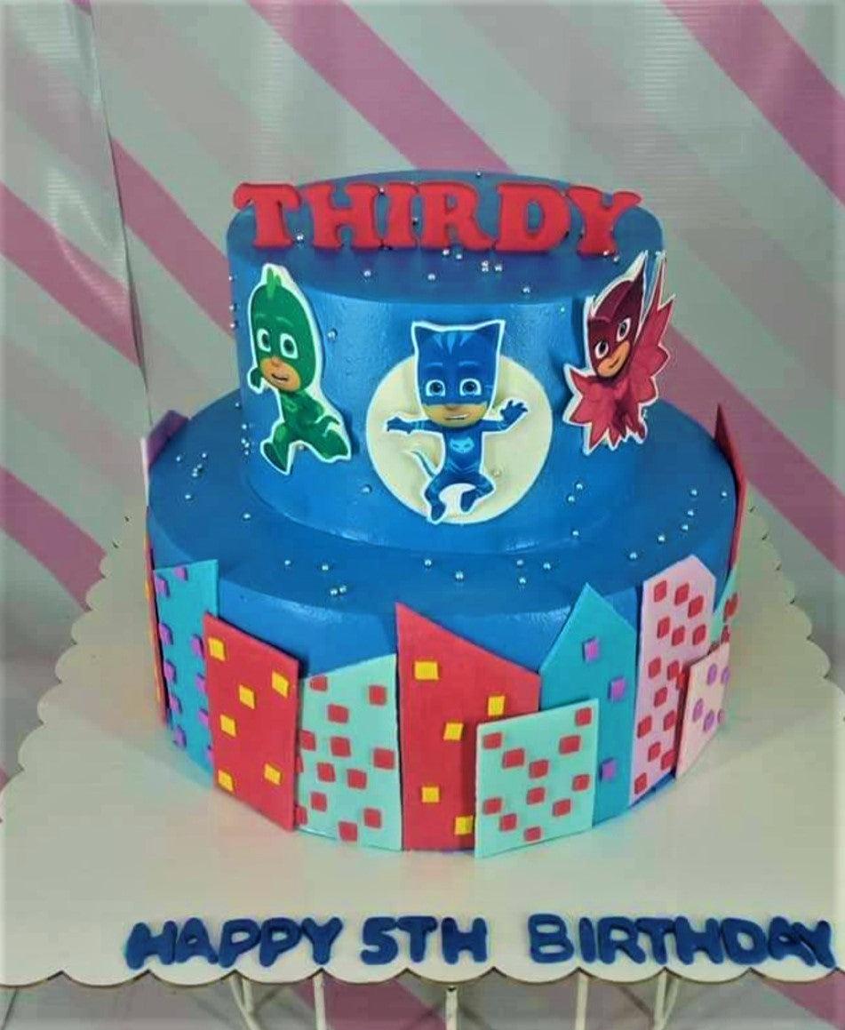 Two-tier blue PJ Masks themed birthday cake with city skyline fondant panels and character printouts.