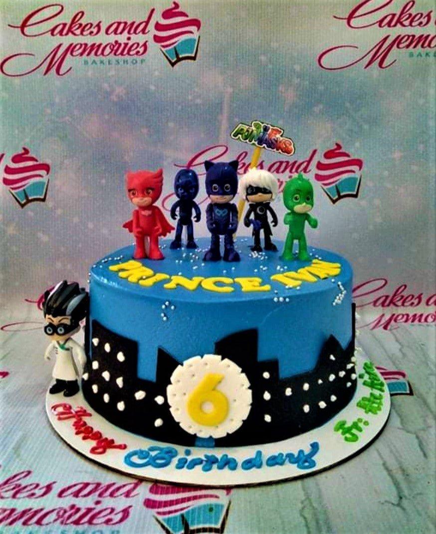 Blue PJ Masks themed birthday cake with character figurines, a black city skyline silhouette, and yellow icing text for Prince Ivan.