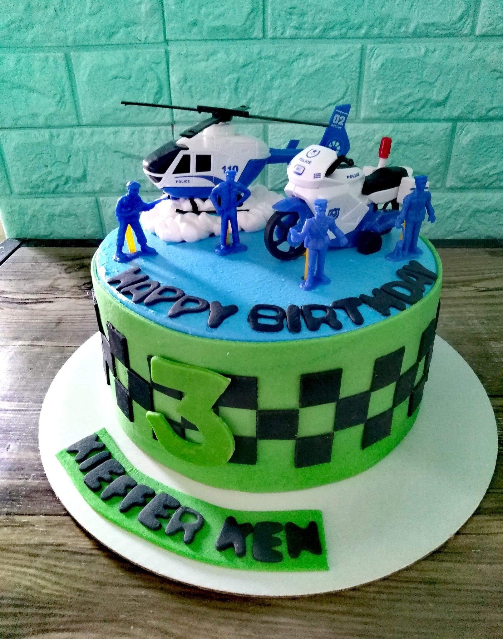 Police themed birthday cake with toy helicopter, motorcycle, and police figurines on blue and green icing with checkered pattern.
