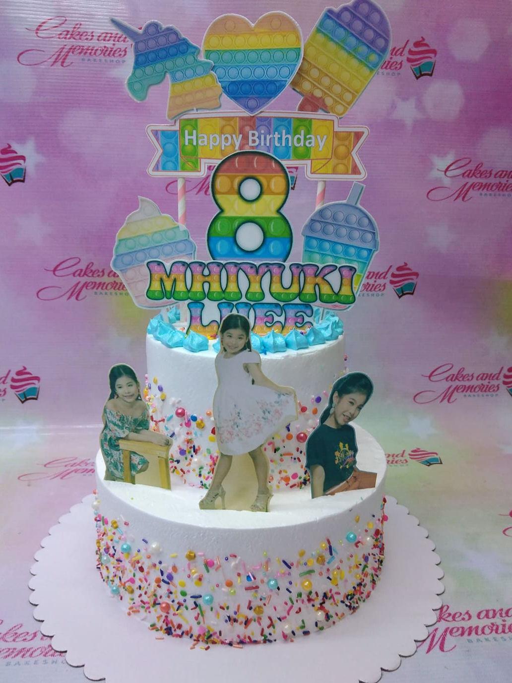 Two-tier white pop-it themed birthday cake with rainbow toppers, colorful sprinkles, and photo printouts of a young girl.