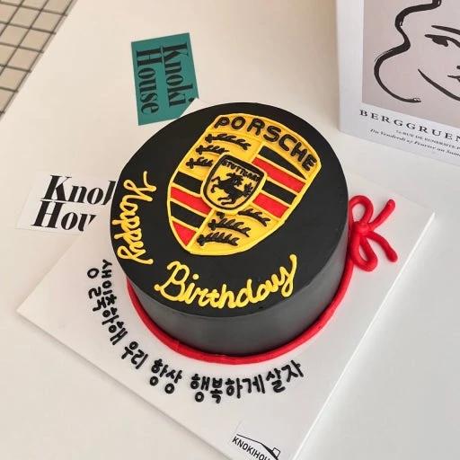 Black minimalist bento cake featuring a yellow and red Porsche logo with yellow piped birthday message and red ribbon detail.