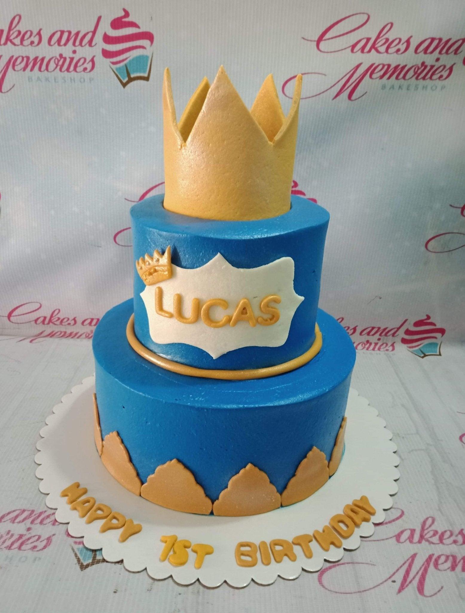 Two-tier blue royal prince cake with a large gold crown topper and gold scalloped fondant accents.
