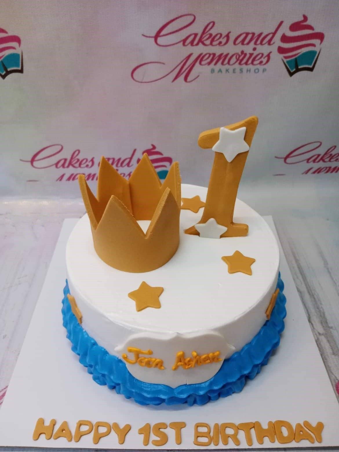 White 1-tier birthday cake with a large gold crown, a gold number one topper, and blue icing ruffles at the base.