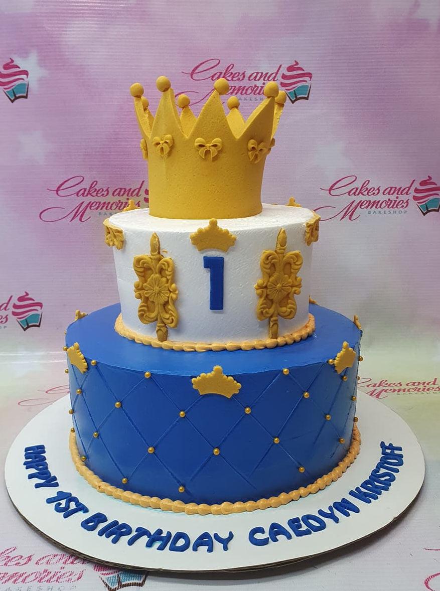 Royal blue and white 2-tier prince birthday cake with a large gold crown topper and quilted icing pattern.