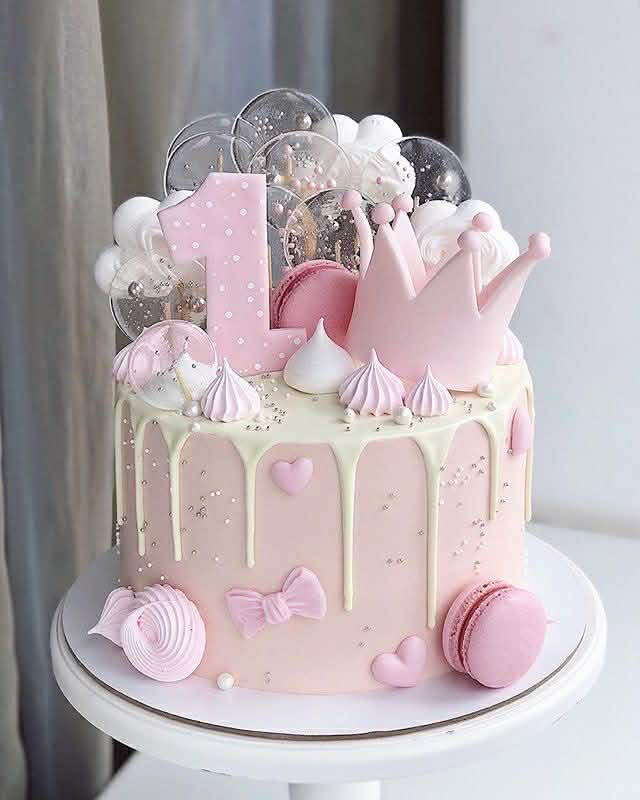 Pink 1st birthday cake with a large pink crown, number one topper, pink macarons, and white chocolate drip.