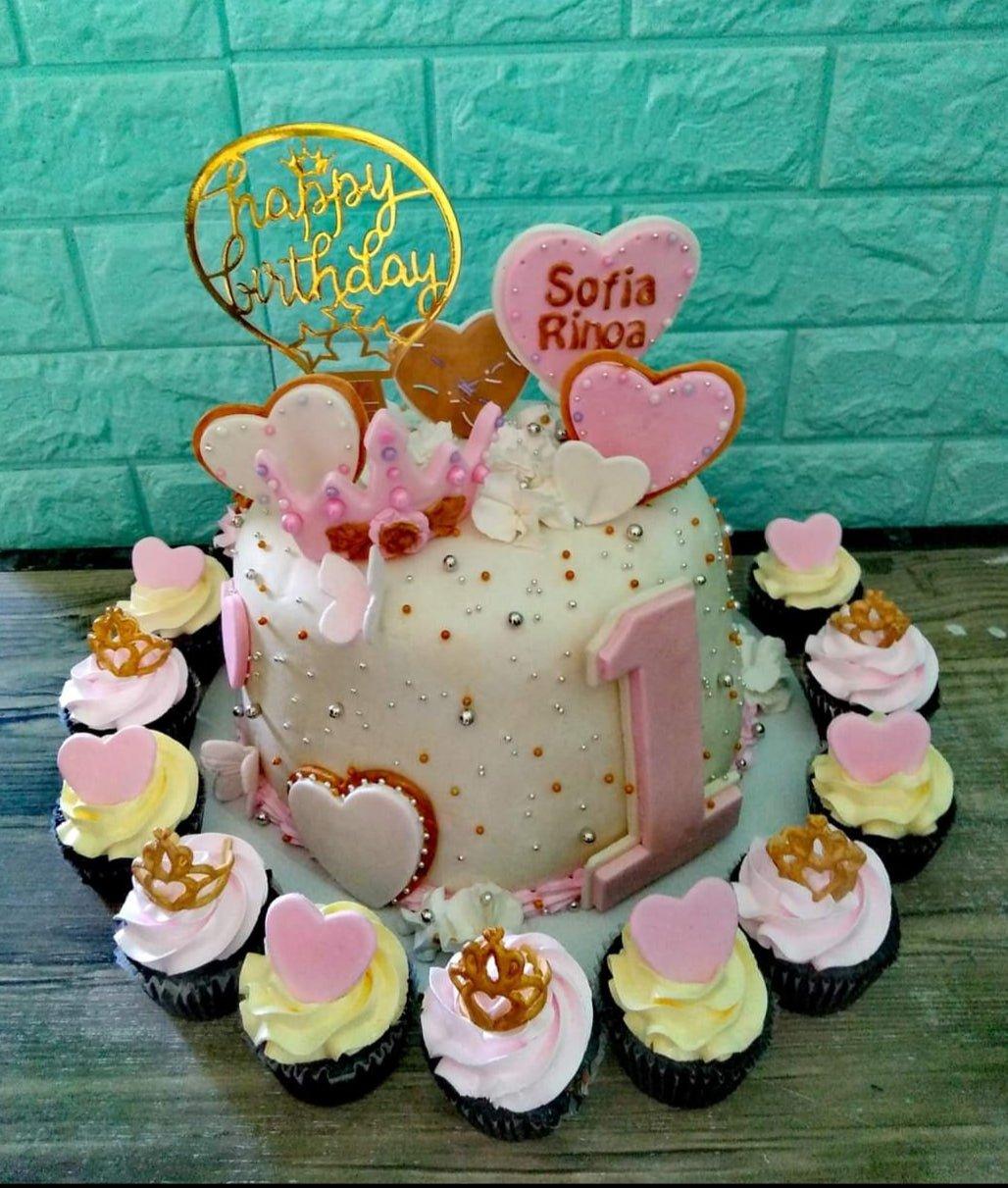 White 1-tier birthday cake with pink heart toppers, a gold crown, and a large pink number one, surrounded by pink and yellow cupcakes.