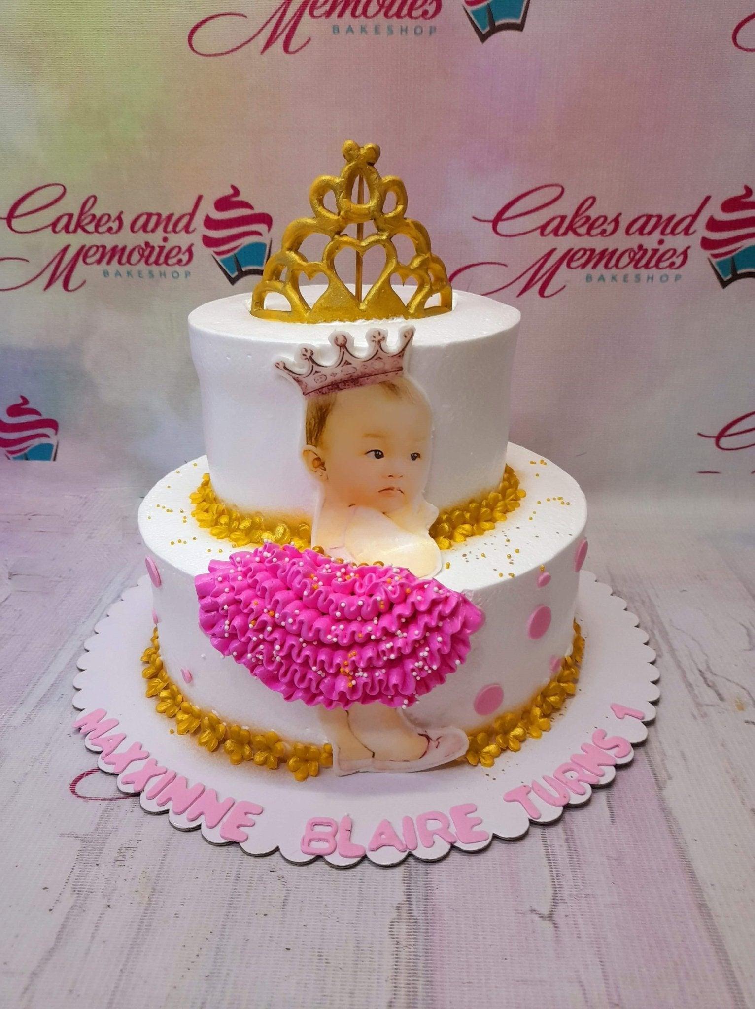 Two-tier white soft-icing cake with a gold crown toy, a baby girl printout topper, and pink ruffled icing decorations.