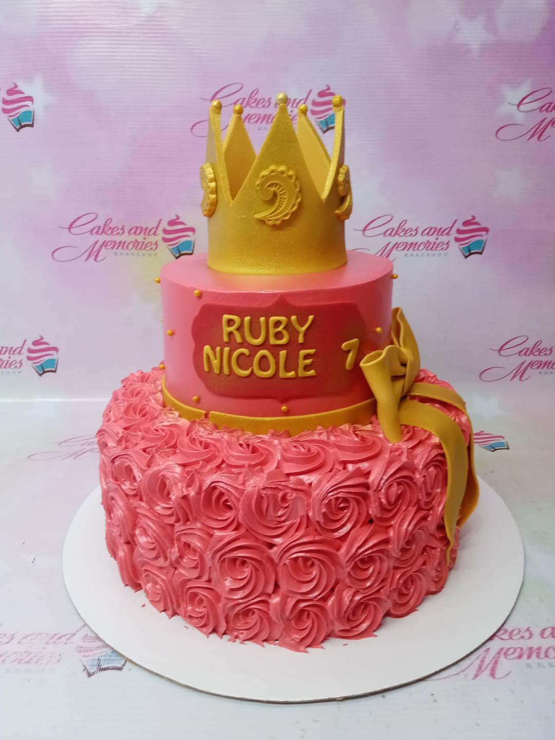 Pink two-tier princess cake with a large gold crown topper, gold ribbon, and pink icing rosettes on the bottom tier.