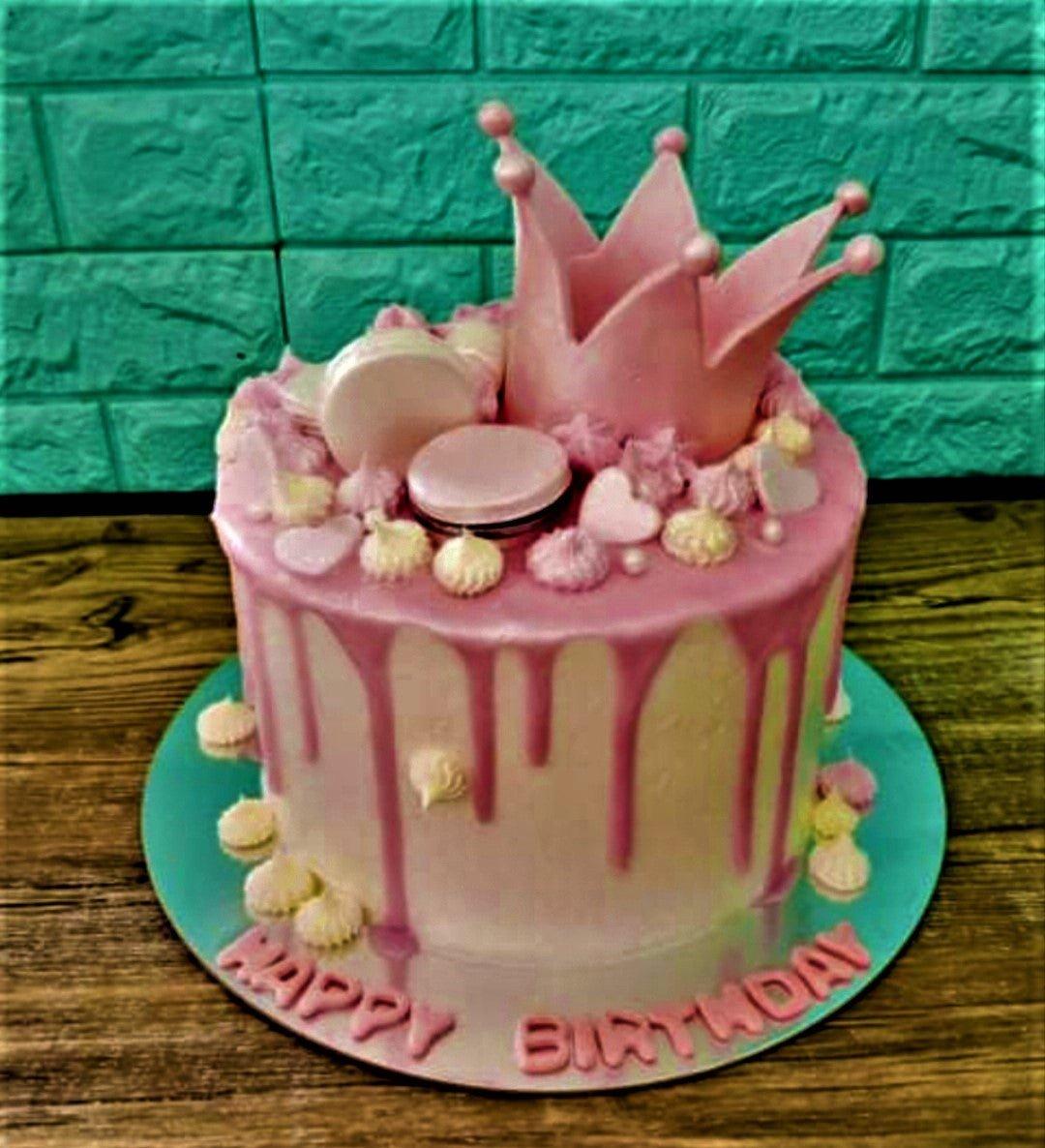 Pink and white princess birthday cake with a pink crown topper, pink drip, macarons, and meringue kisses.