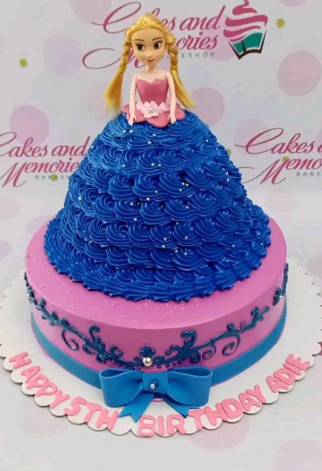 Princess doll cake with a voluminous blue icing dress on a pink base tier with blue scrollwork and a large blue bow.