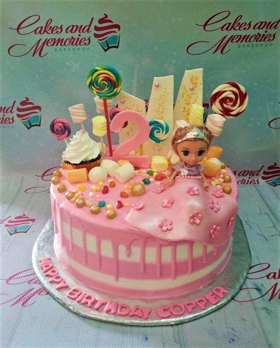 Pink and white striped birthday cake with a princess doll, lollipops, marshmallows, and a pink number 2 topper.