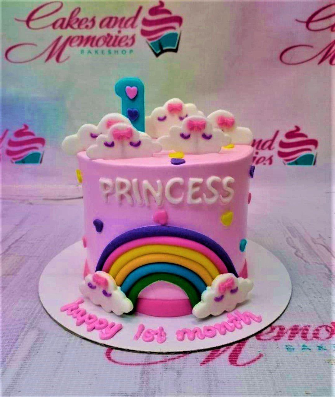 Pink princess themed cake with a fondant rainbow, clouds with bows, and a blue number one topper for a first month celebration.