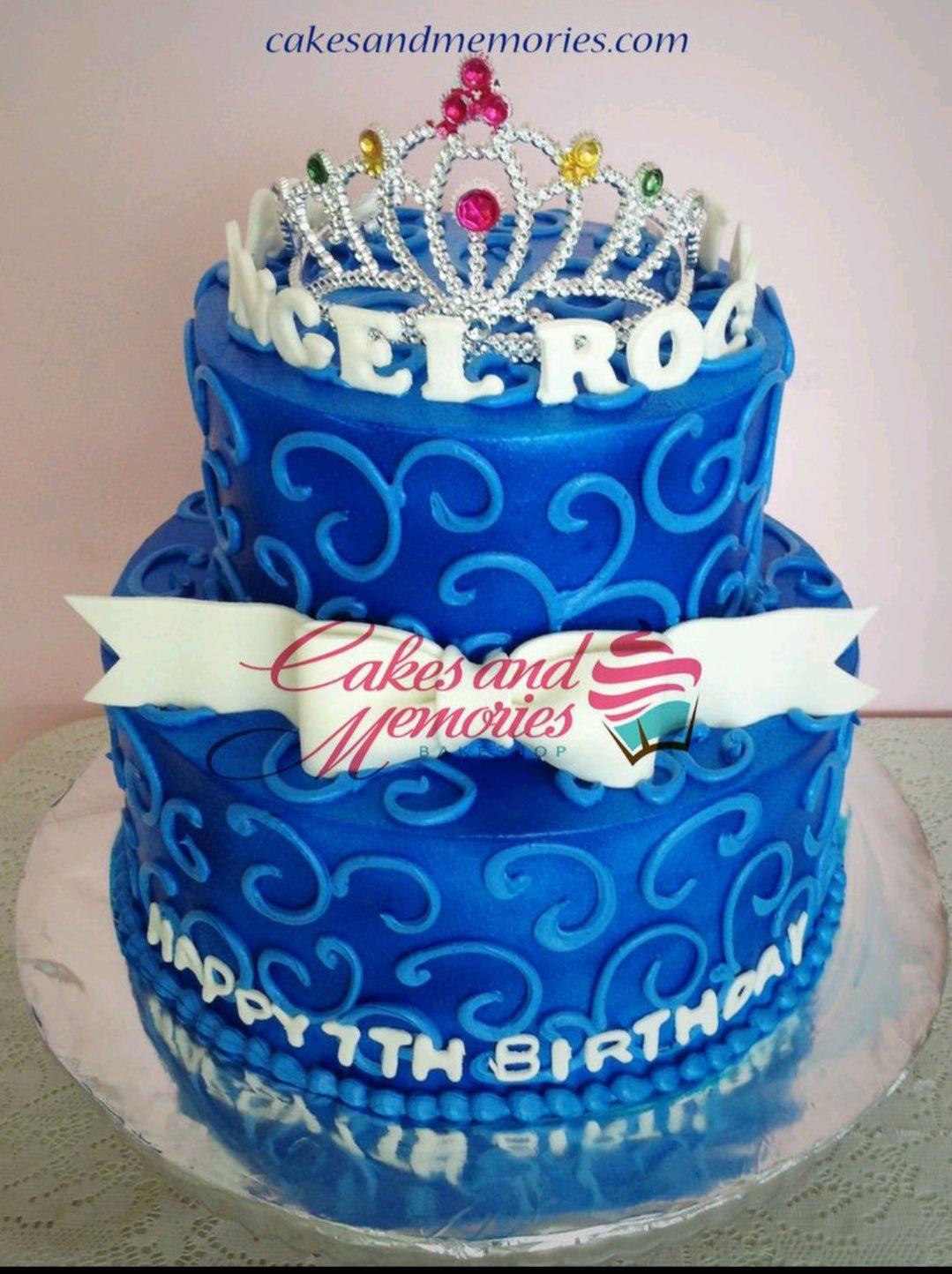 Two-tier royal blue fondant cake with intricate light blue icing swirls, a white fondant bow, and a silver rhinestone tiara on top.