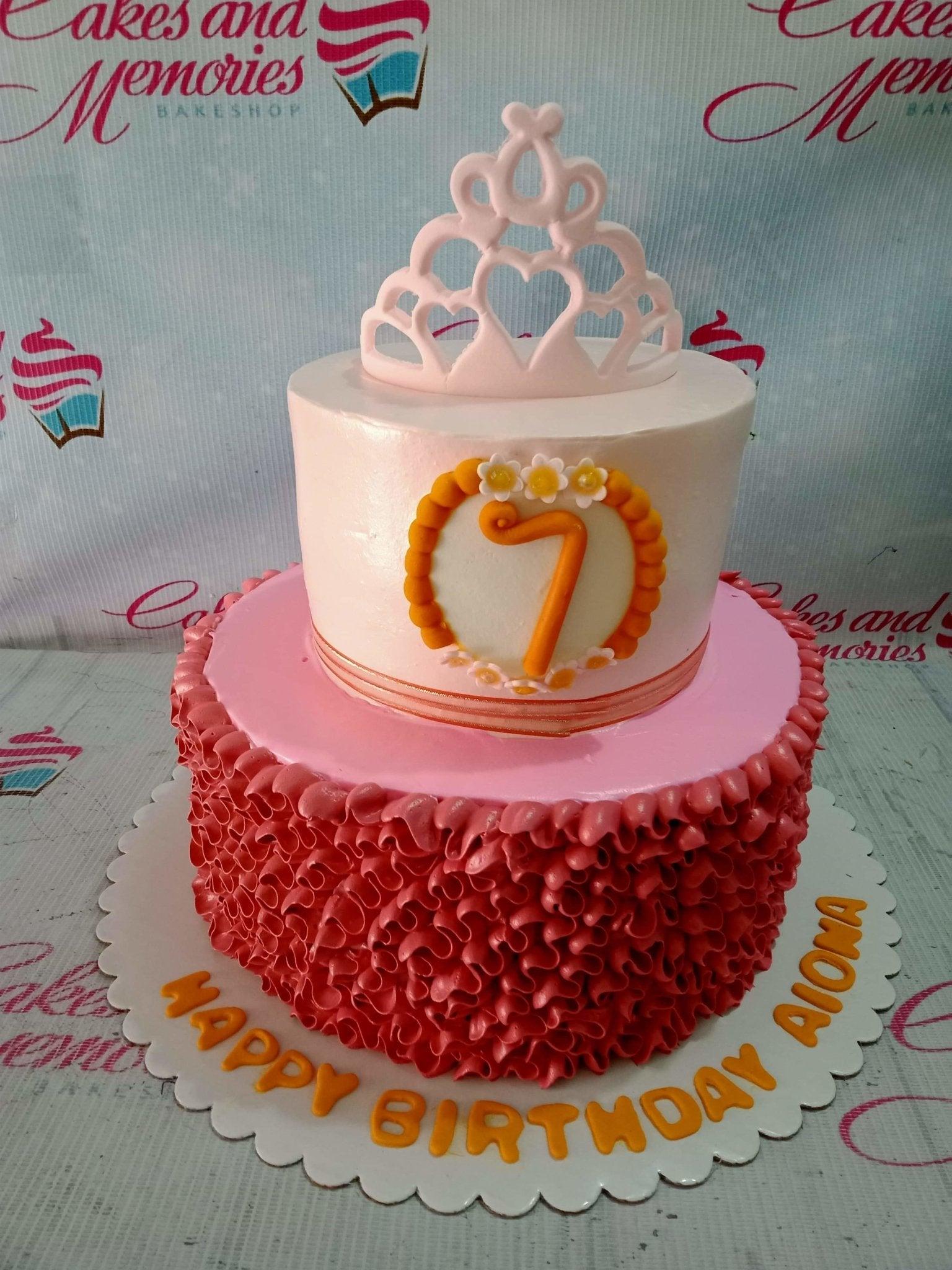 Two-tier pink and white princess cake with a pink tiara topper, orange number 7, and ruffled icing on the bottom tier.